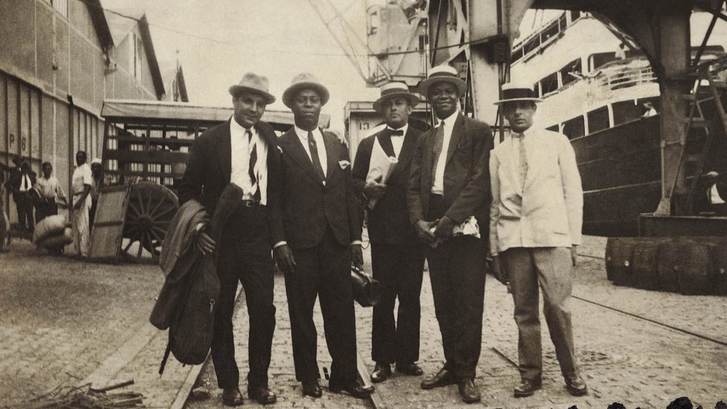 Duque (o primeiro da esquerda para a direita), Donga (o segundo), Pixinguinha (o quarto) e duas pessoas não identificadas, no porto do Rio de Janeiro, possivelmente no embarque dos Oito Batutas para Paris, em 29 de janeiro de 1922. Coleção Pixinguinha/Acervo IMS